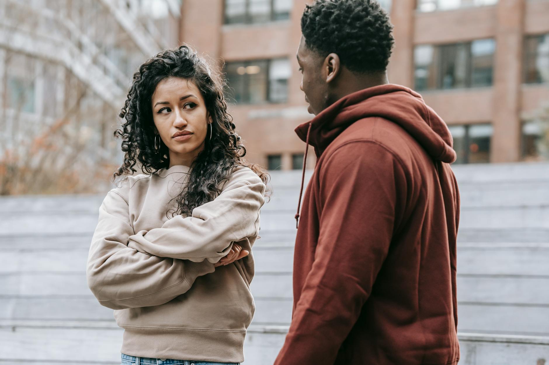 Unset ethnic woman with crossed arms and African American man looking at each other while having quarrel on street with building on blurred background - spring allergy irritability