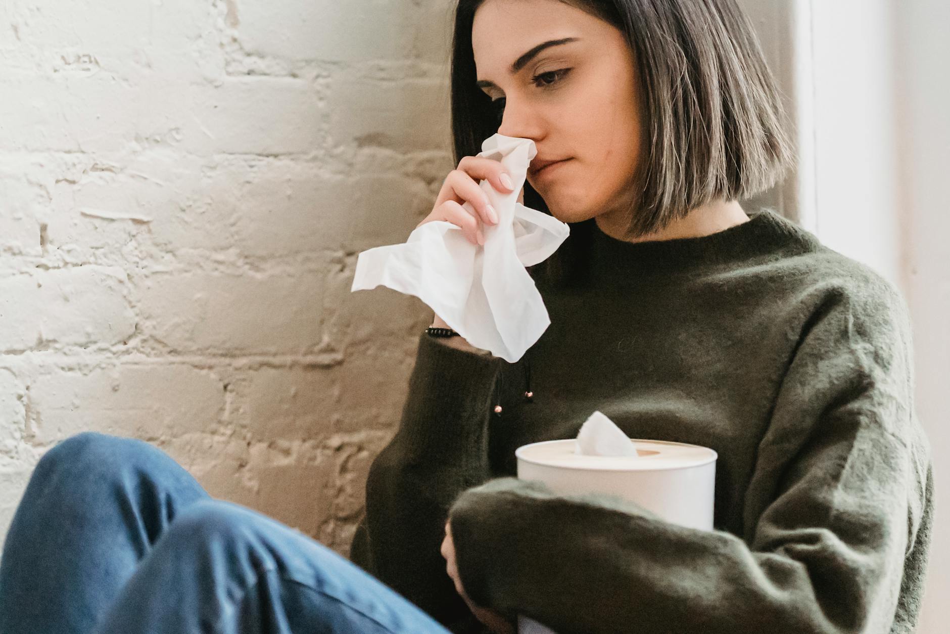 Crop young sad female with dark hair in sweater with tissue in hand crying and looking away sitting near white brick wall at home - spring sad symptoms