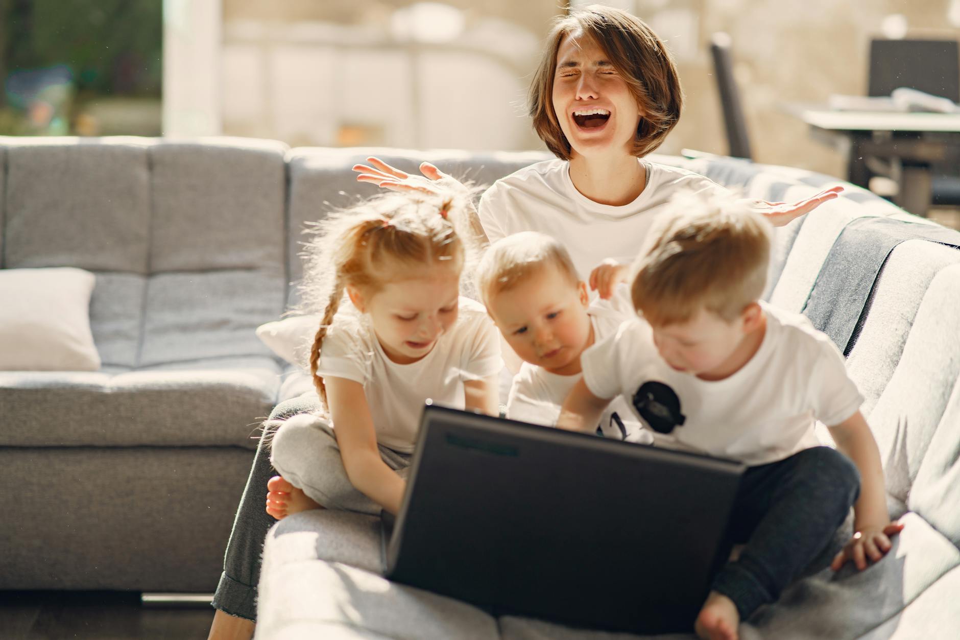 Stressed woman asking for help while sitting with small children playing on laptop together on sofa in living room - spring stress families