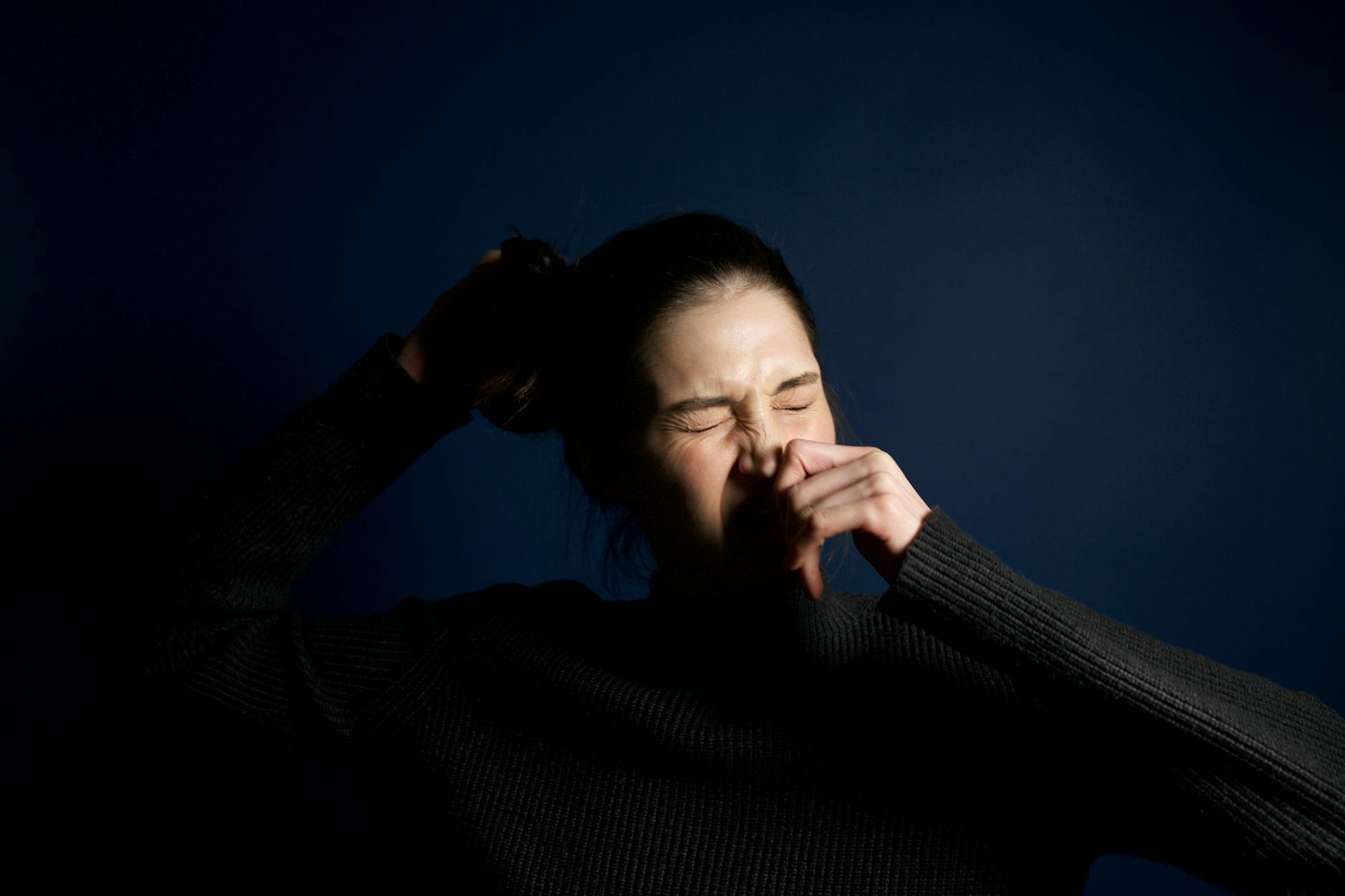 A woman in a dark sweater sneezes against a solid blue background, capturing a moment of candid expression. - teen spring allergies