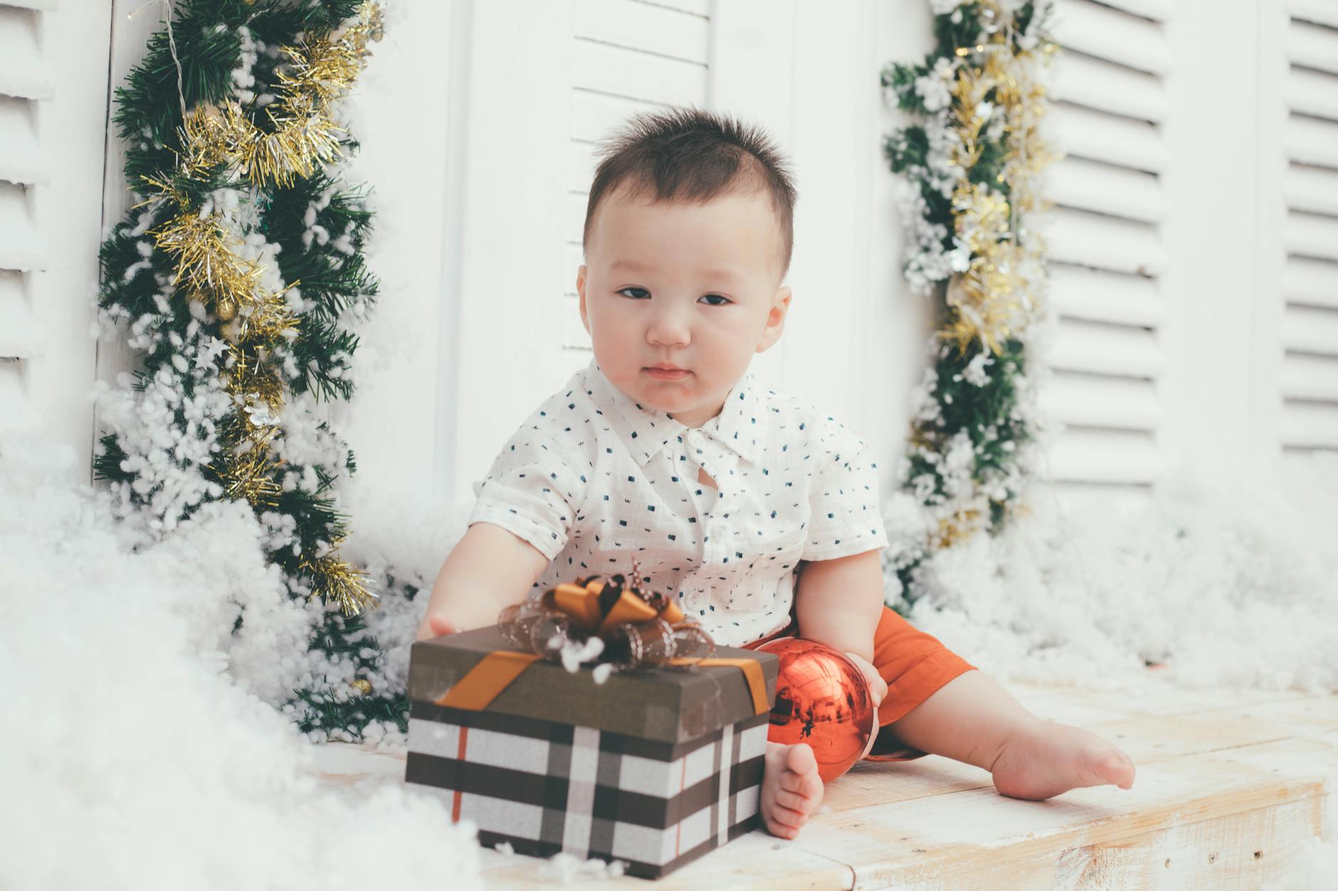 Adorable baby sitting indoors surrounded by festive Christmas decorations, holding a gift box. - toddler independence winter