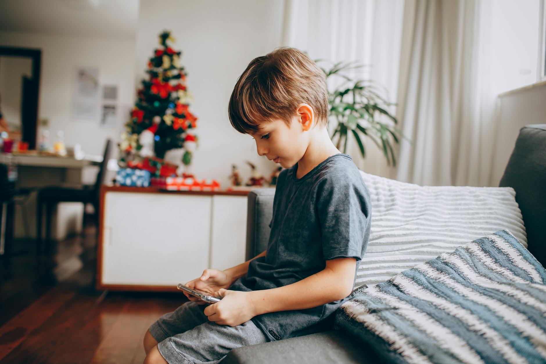 A young boy focusing on his smartphone while sitting near a decorated Christmas tree indoors. - toddler spring tantrums