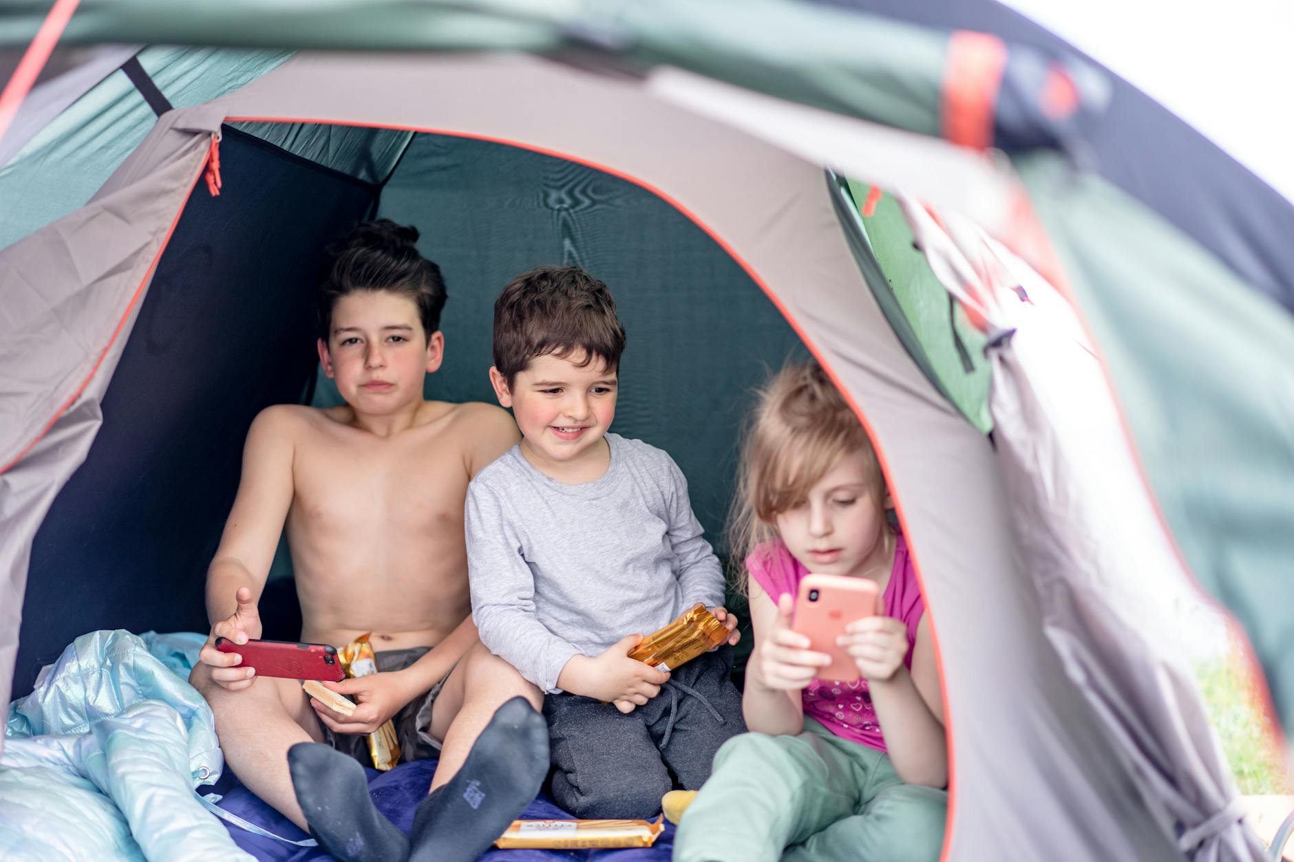 Three children relax in a tent using smartphones during a camping trip, enjoying leisure and snacks. - winter sleep children