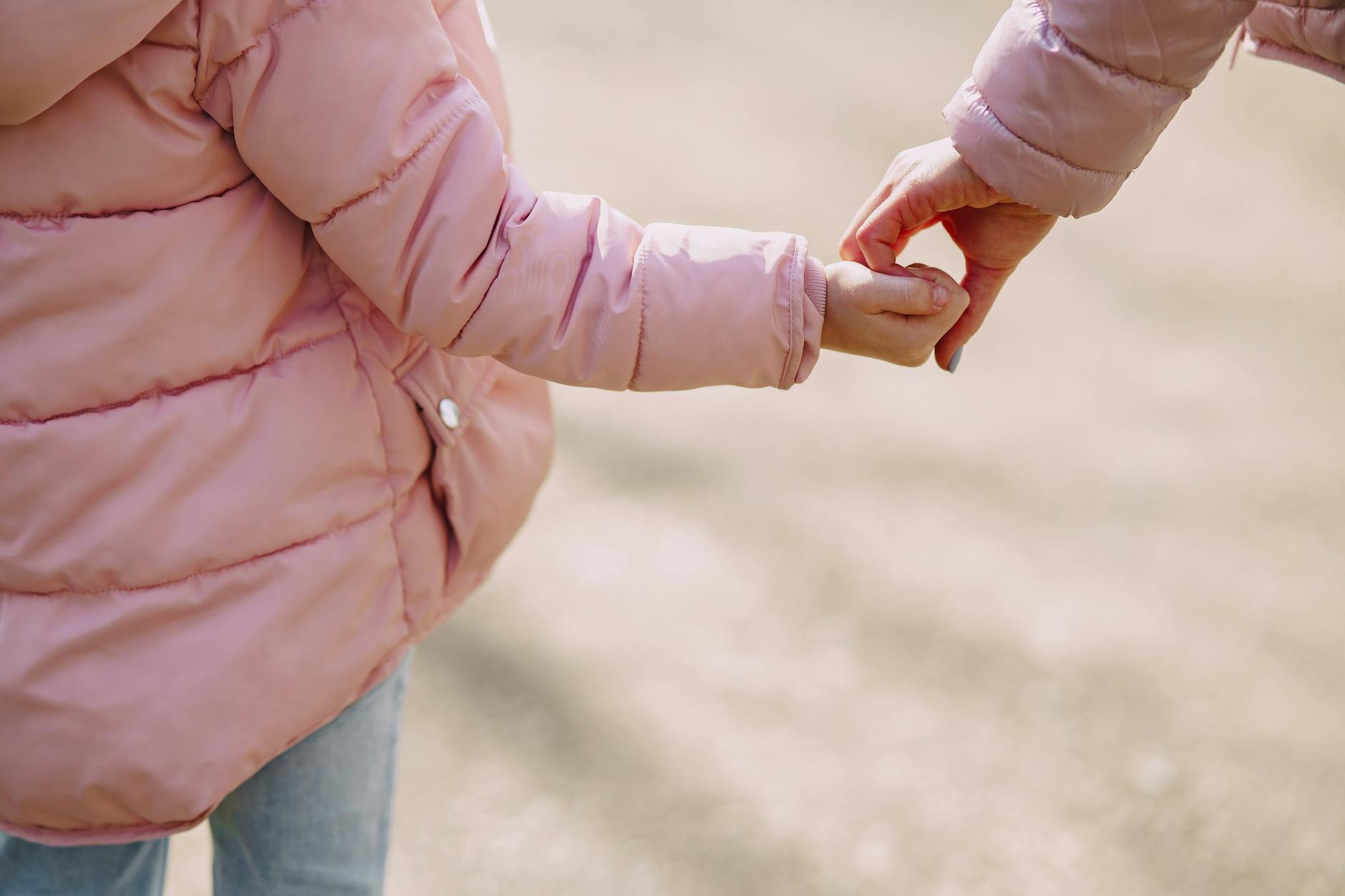 Back view of unrecognizable little girl wearing warm pink jacket holding hands with crop young woman while spending time together in city outside illustrating mother care and support concept - co-parenting after divorce