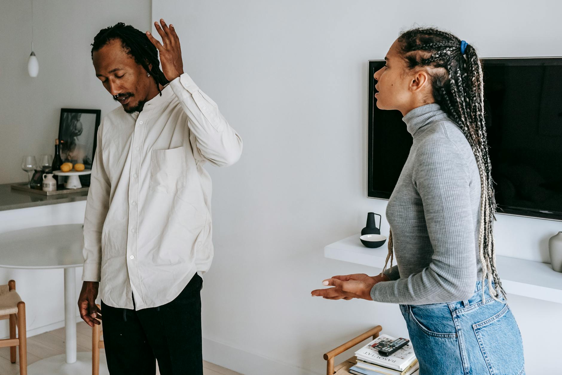 Tired young ethnic man with dreadlocks in casual outfit gesticulating while having argument with wife at home - couple conflict resolution