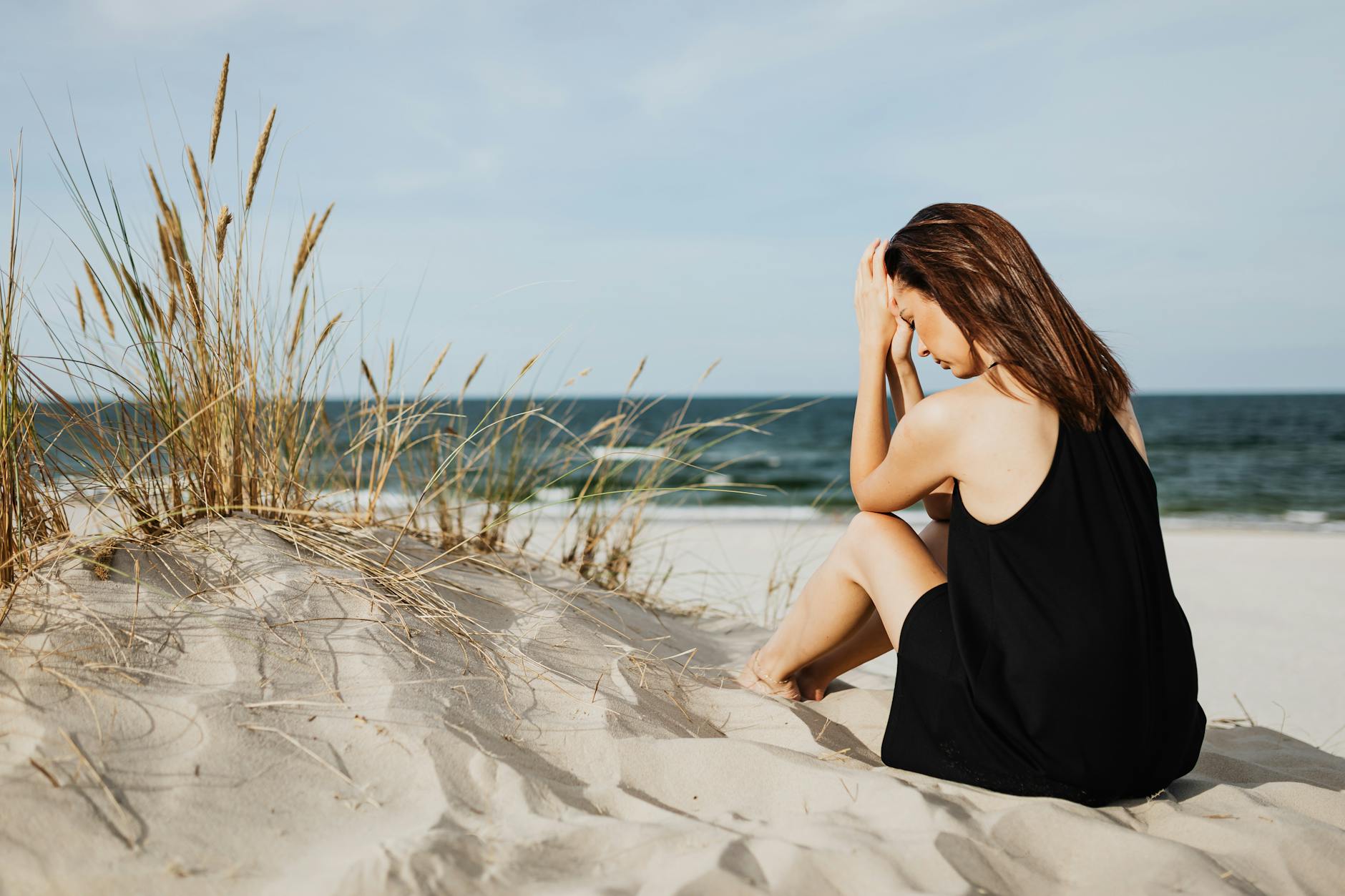 A woman in a black dress sits on sandy dunes by the ocean, thoughtful and serene. - letting go resentment