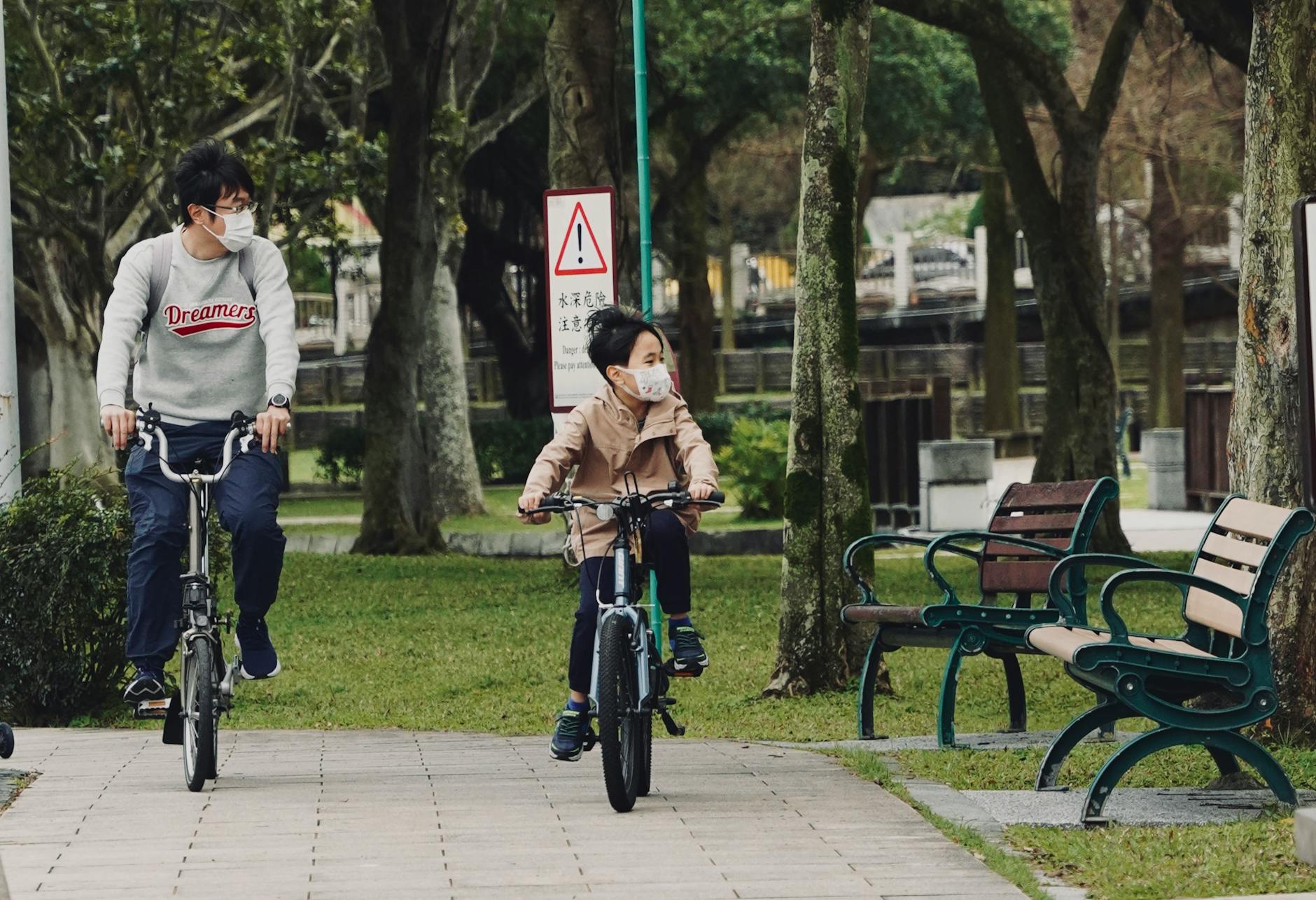 Father and son riding bicycles in the park, both wearing masks for safety. - post pandemic family