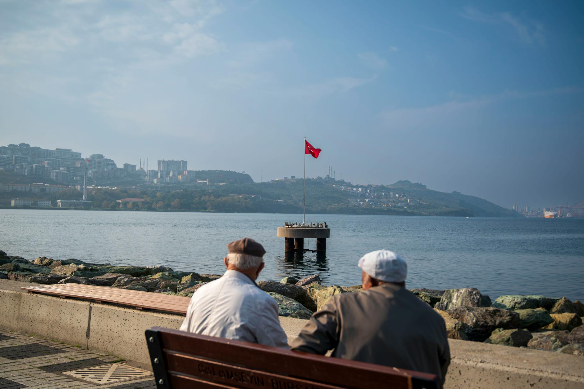 Two elderly men sit on a bench by the sea, viewing a distant flag over calm waters. - red flags relationships