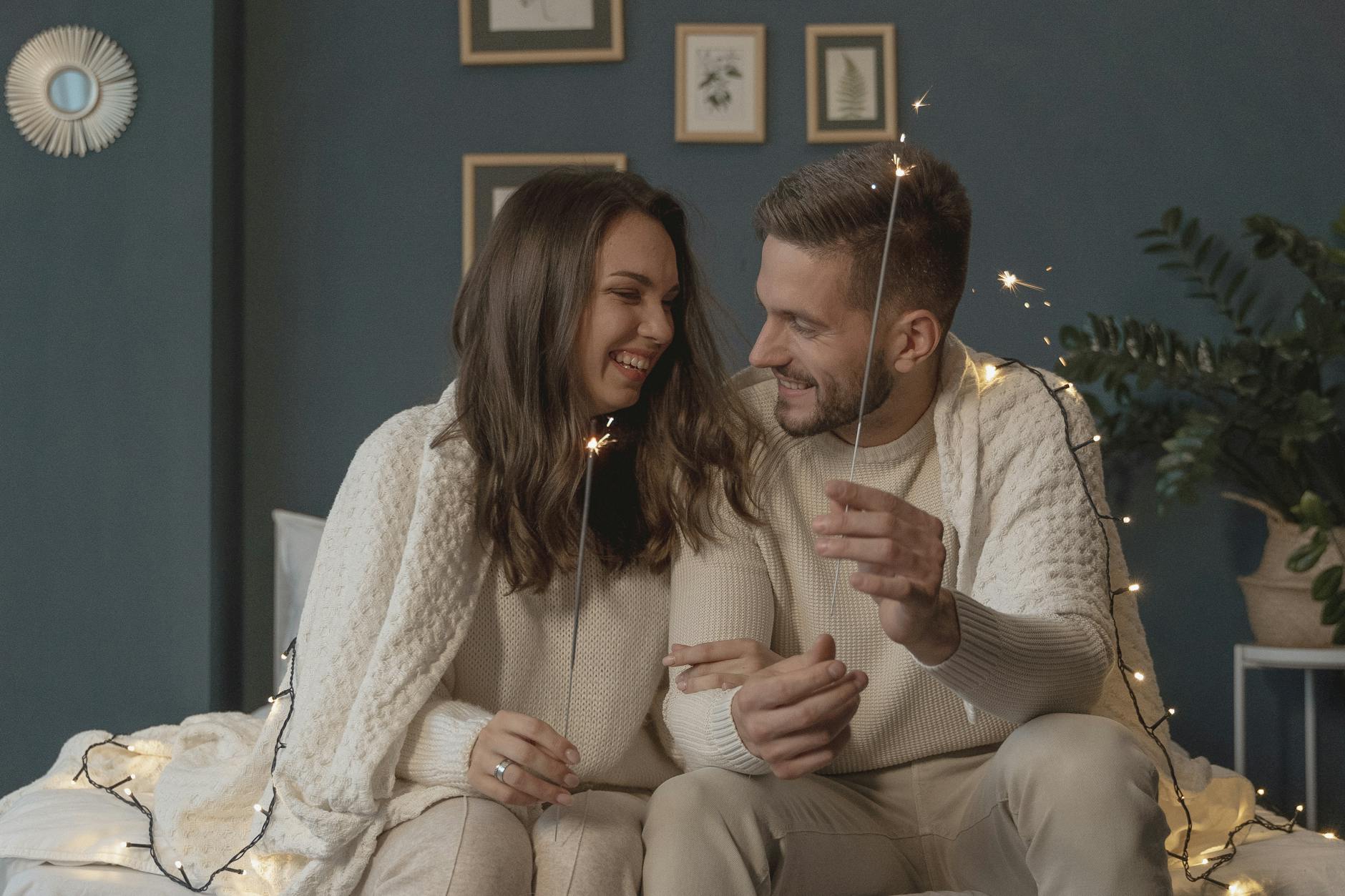 A loving couple smiles warmly while playing with sparklers in a cozy bedroom setting. - reignite relationship spark