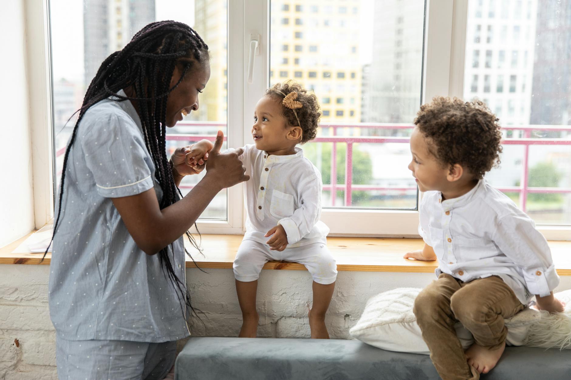 A mother and her young children enjoying playful interaction on a windowsill indoors. - sibling rivalry tips