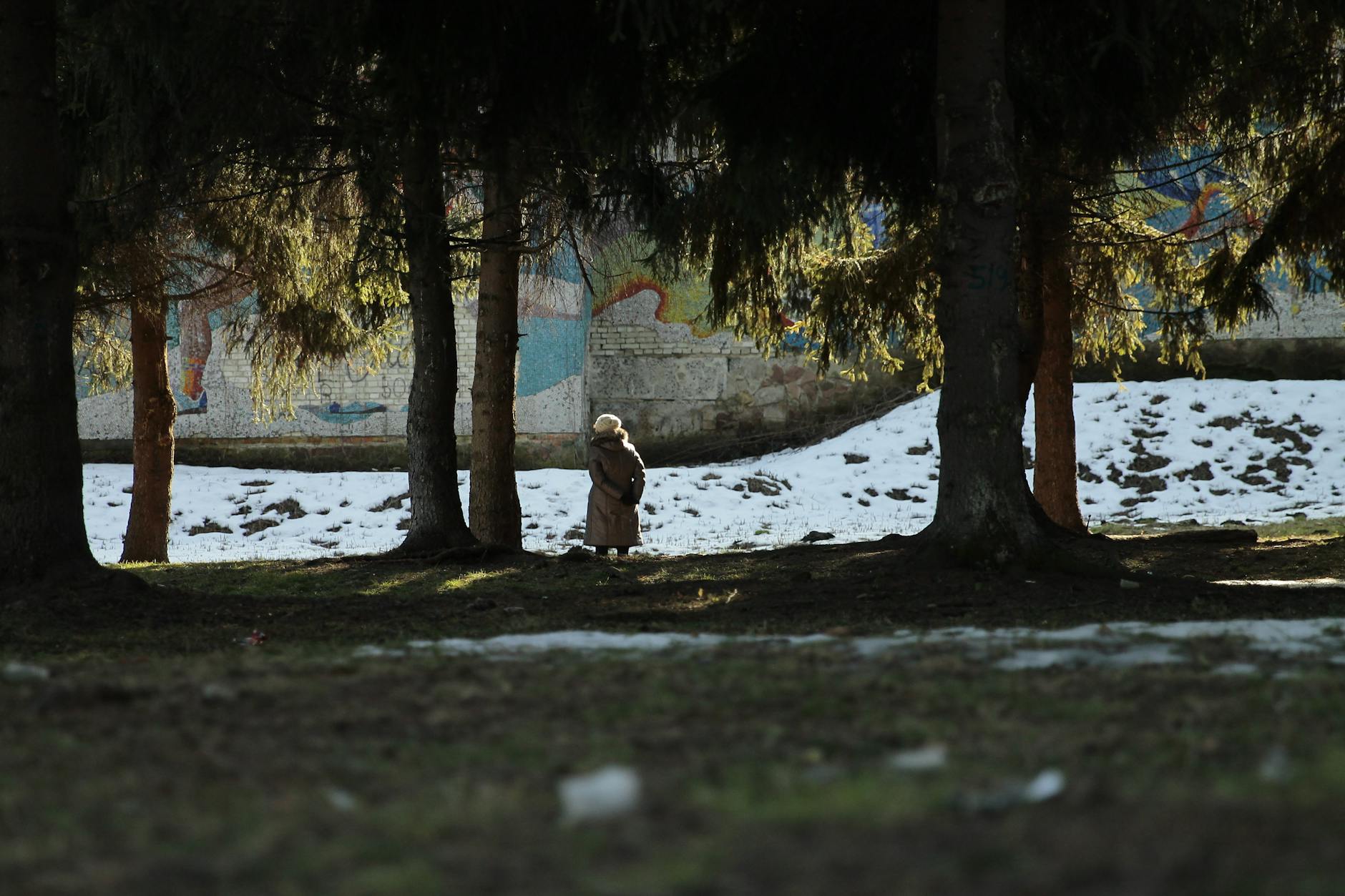 A woman in a coat walks among trees near a snowy field with a mural wall. - spring loneliness