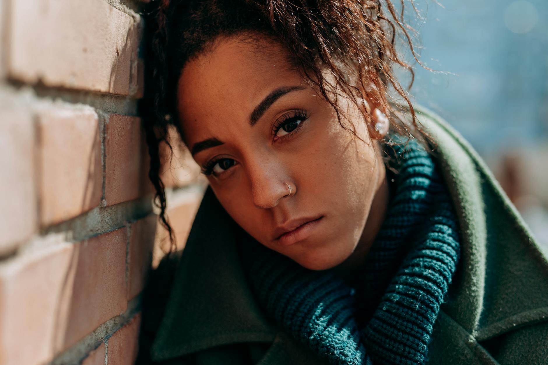 A thoughtful woman in a green coat leans against a brick wall on a sunny day. - spring sad coping
