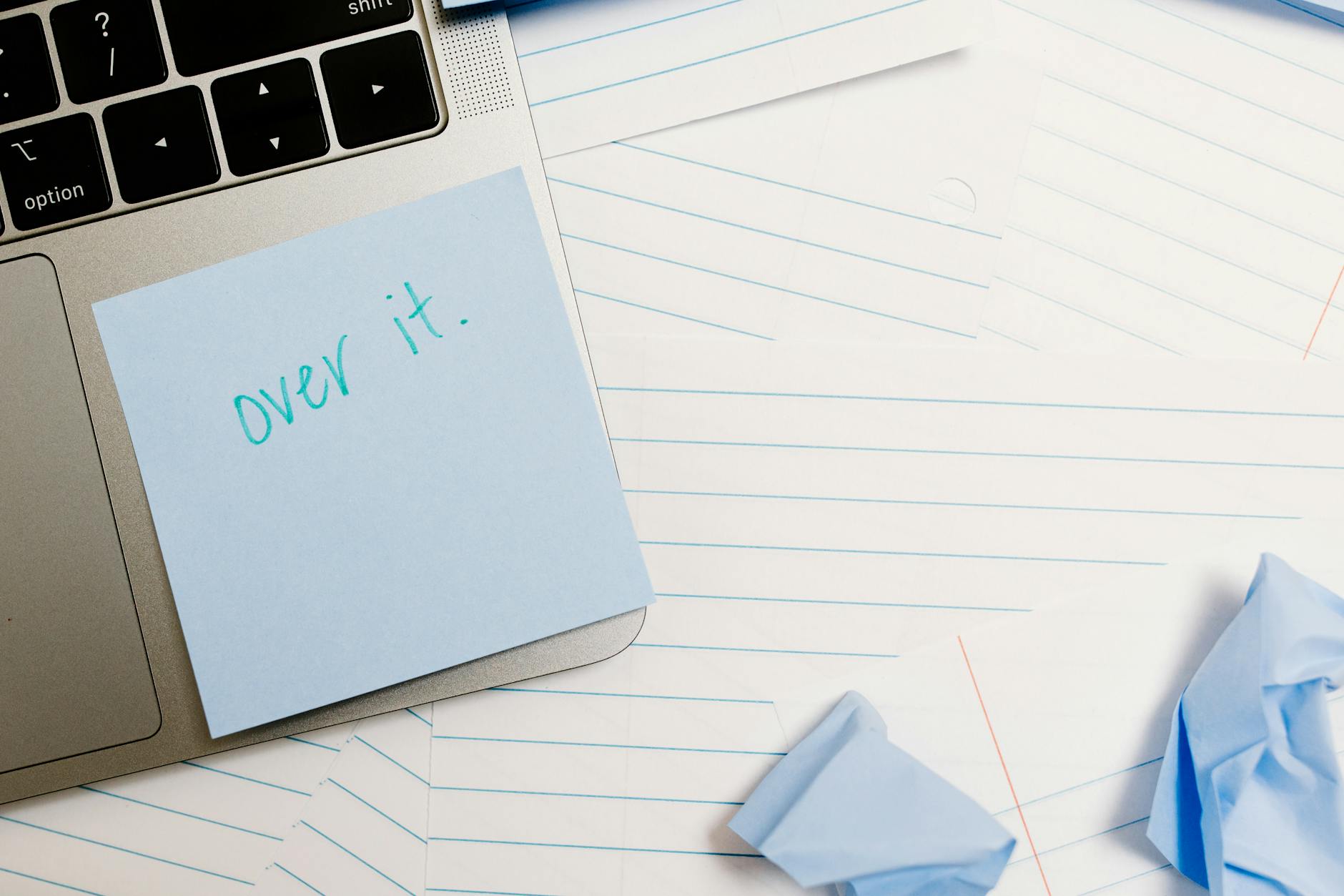 Close-up of laptop with sticky note saying 'over it' surrounded by lined papers and crumpled notes. - stop overthinking