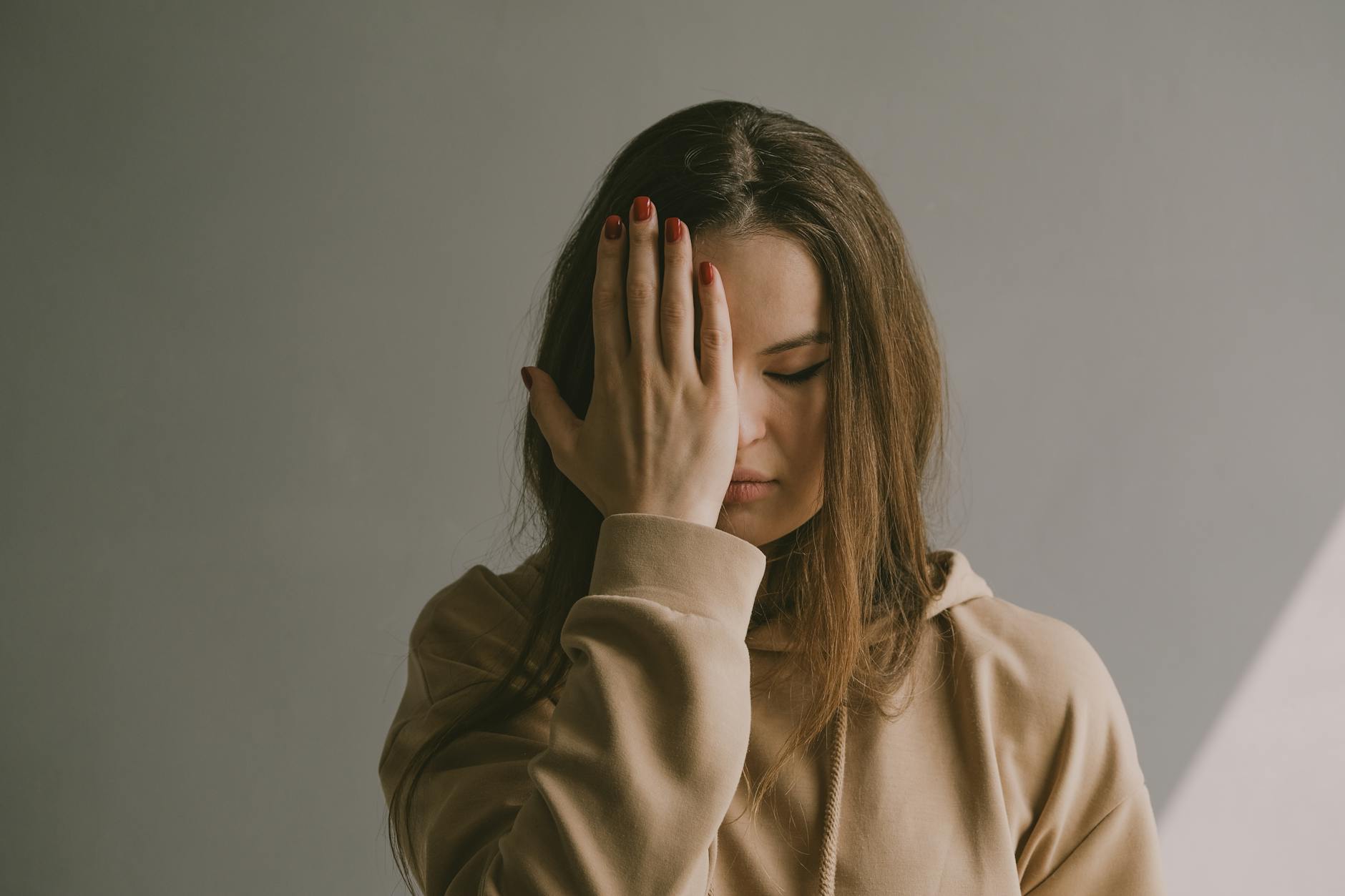 A woman with brown hair wearing a hoodie covers her face with her hand, depicting emotion. - stop overthinking
