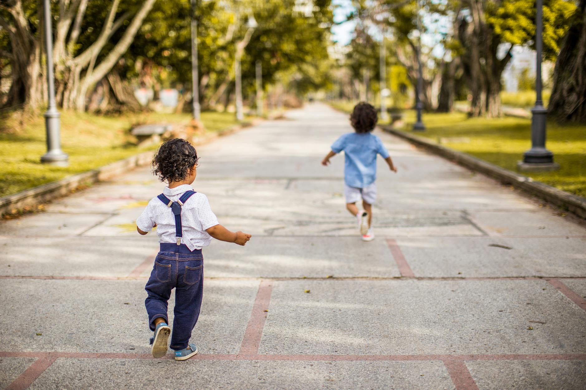 Two young children running joyfully on a paved pathway in a scenic park setting, enjoying a sunny day. - toddler outdoor activities