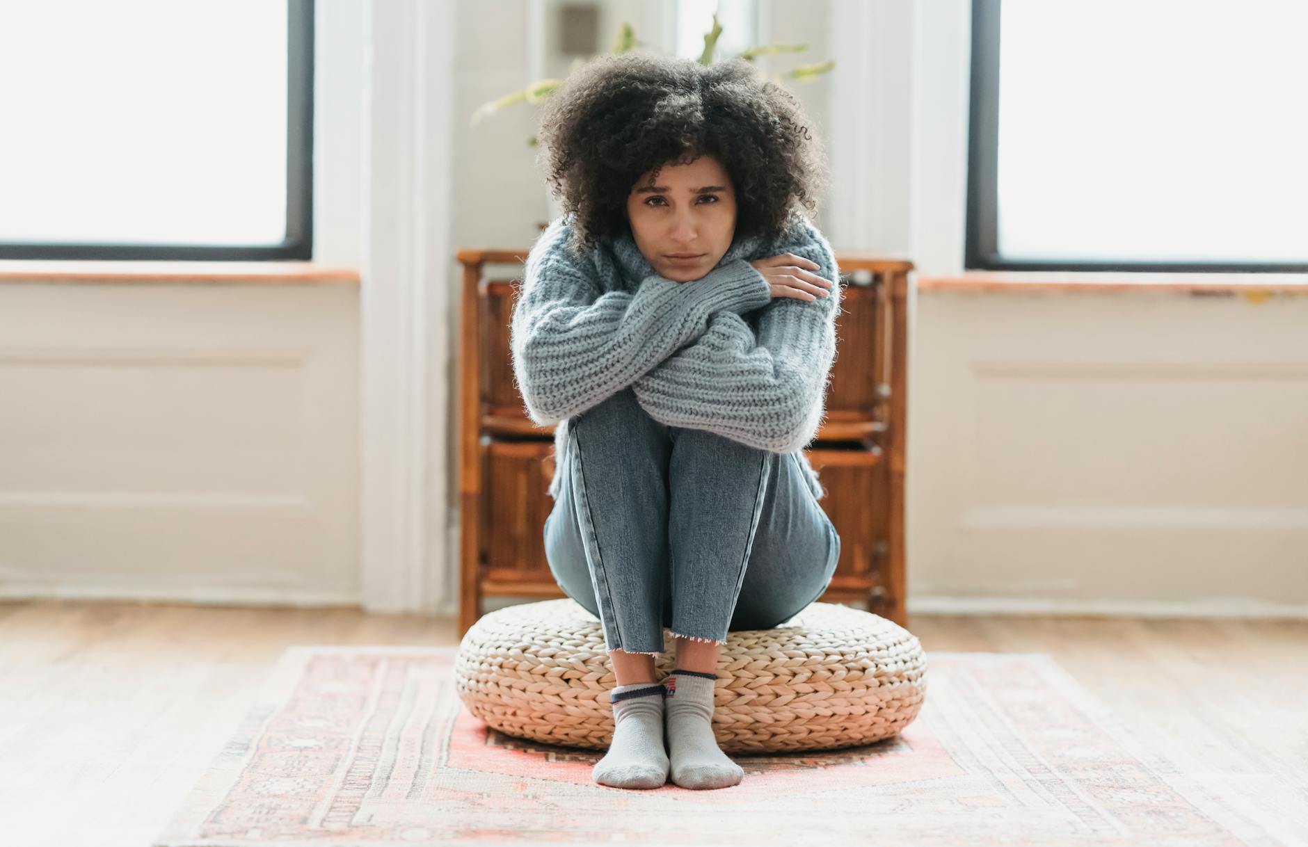 Full body of sad African American female looking at camera while sitting on rattan mat in light room at home - cbt for spring sad