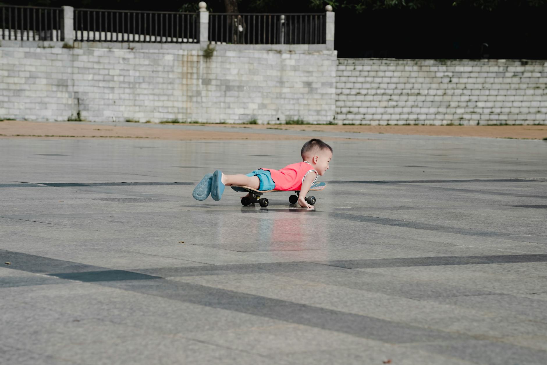 A young child in a red shirt lays flat on a skateboard in an outdoor paved area. - independent outdoor play