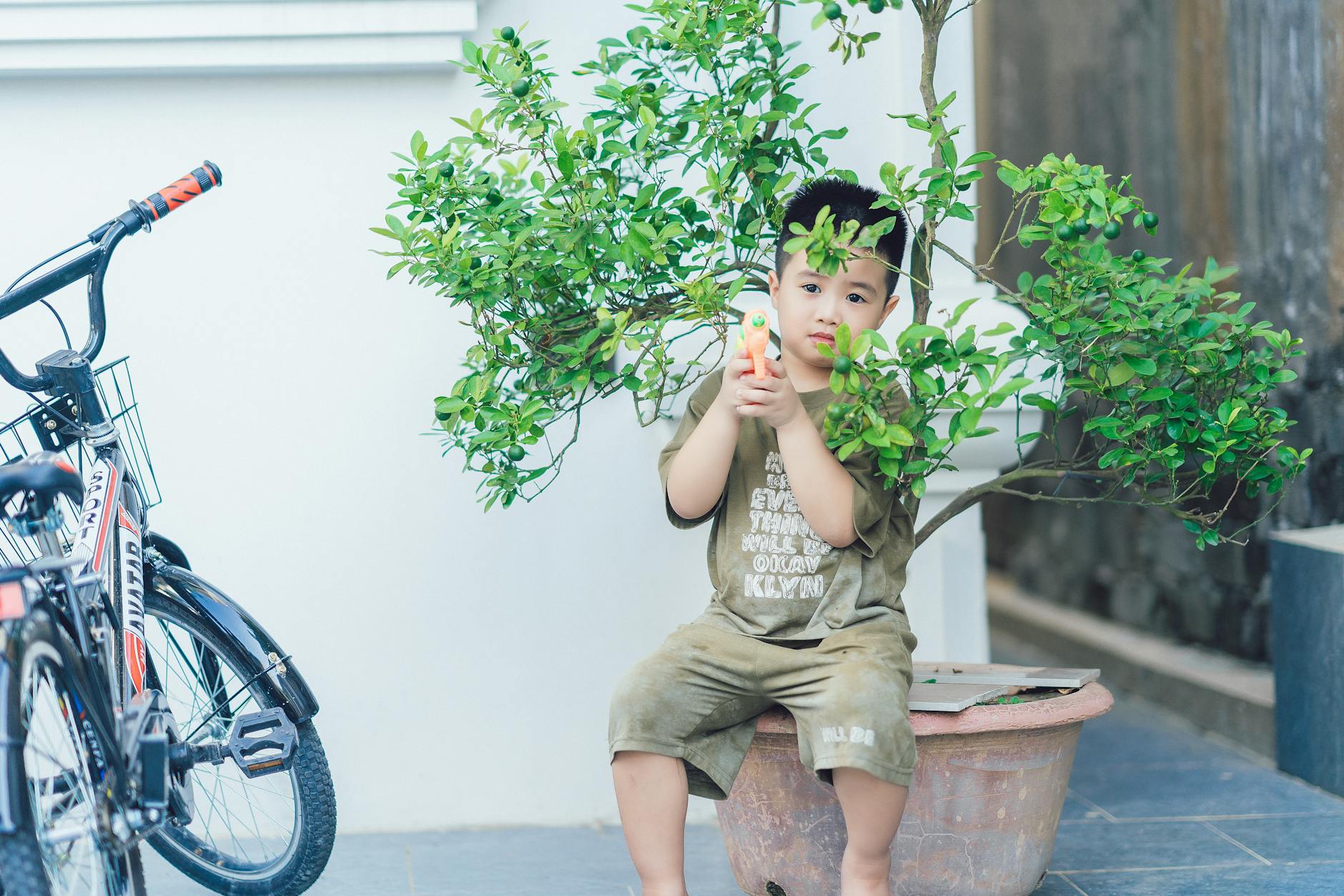 A young boy with a toy gun sits by a potted plant and bicycle outdoors. - independent outdoor play