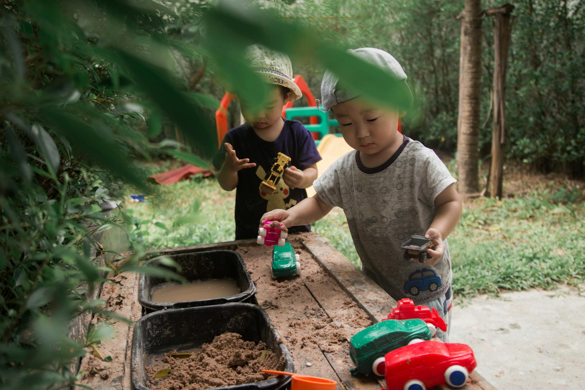 Young children engaged in imaginative play with toy cars in a garden sandbox. - independent outdoor play