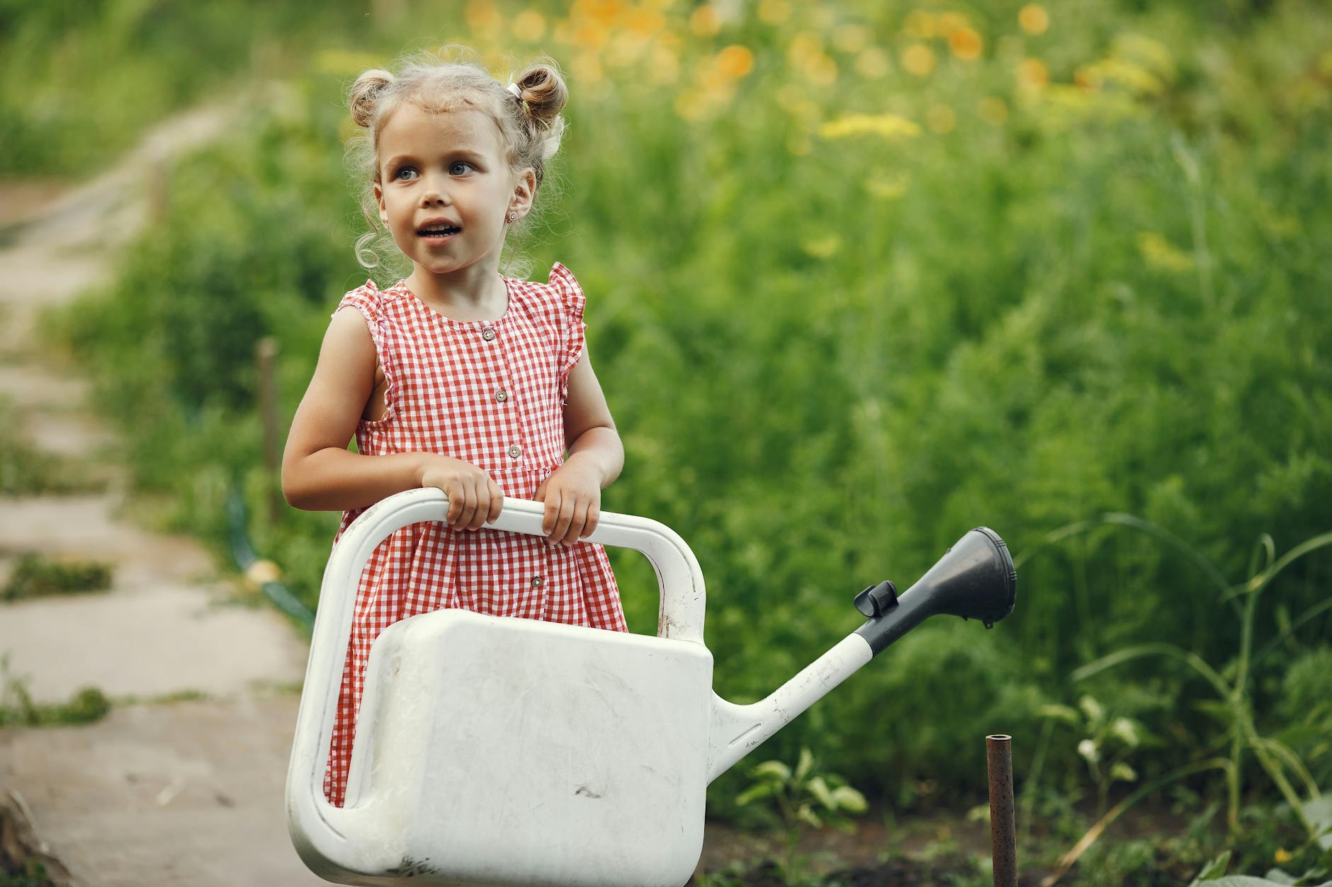 A young girl in a red checkered dress holding a watering can in a lush summer garden. - preschool garden activities