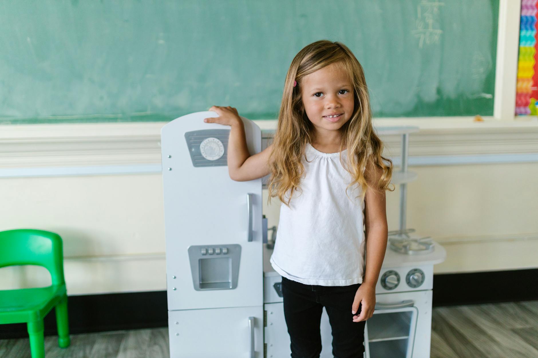 A cheerful young girl stands in a classroom near a play kitchen, showcasing a lively learning environment. - preschooler self identity