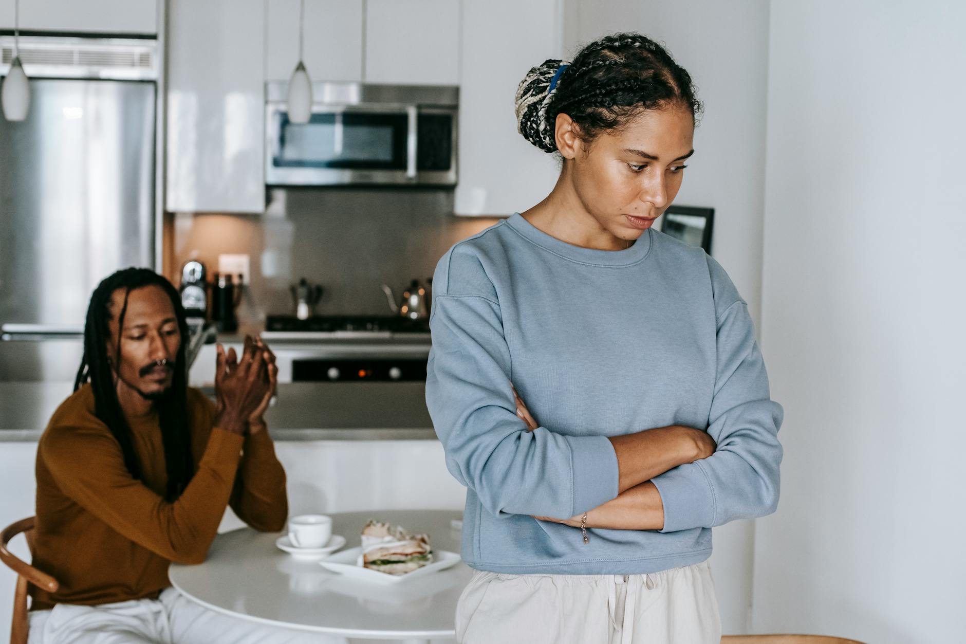 Young discontented African American female with folded arms against male partner at kitchen table during quarrel - relationship conflict spring