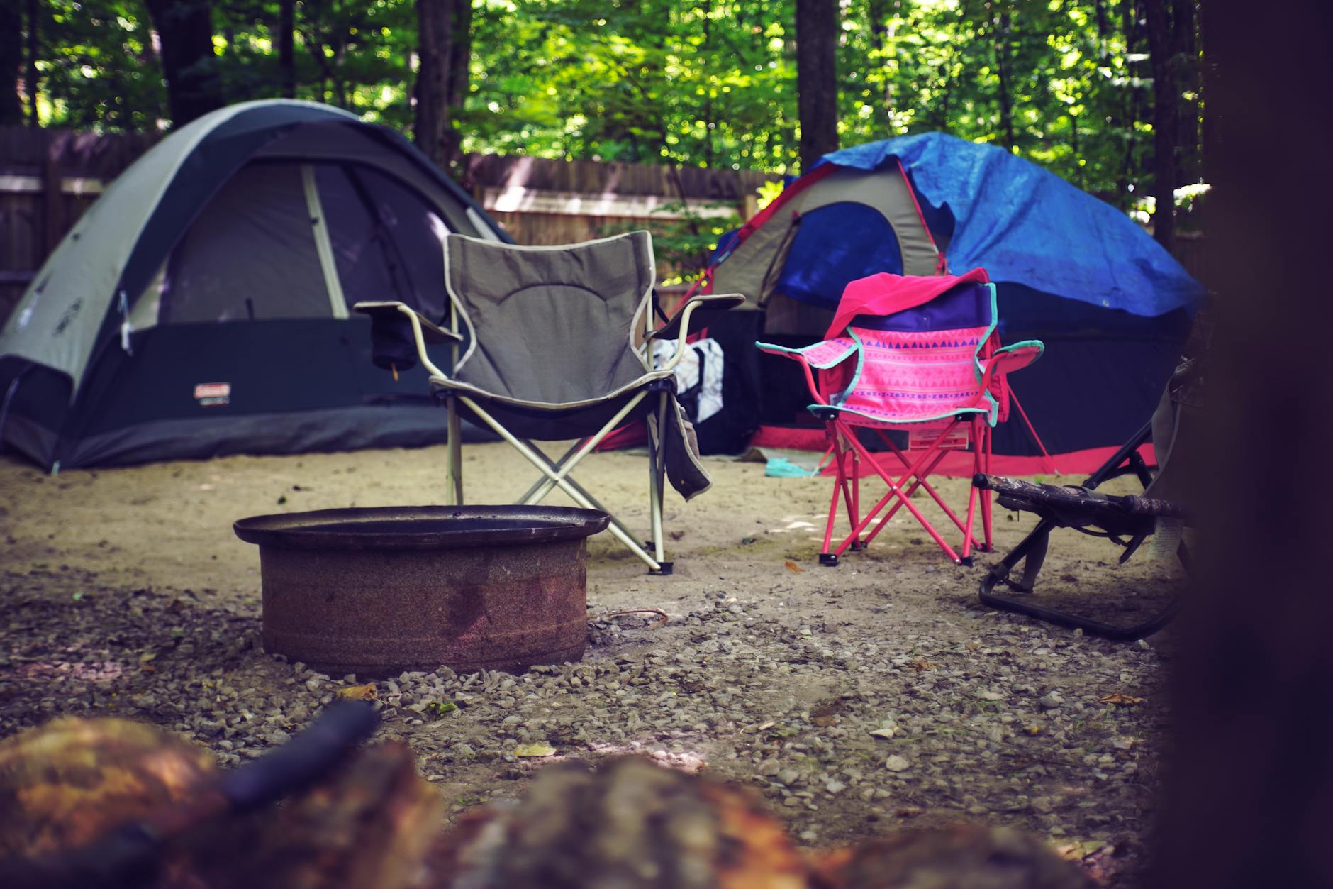 A serene campsite in the woods with colorful tents, camping chairs, and a fire pit, perfect for a leisure getaway. - summer camp fears