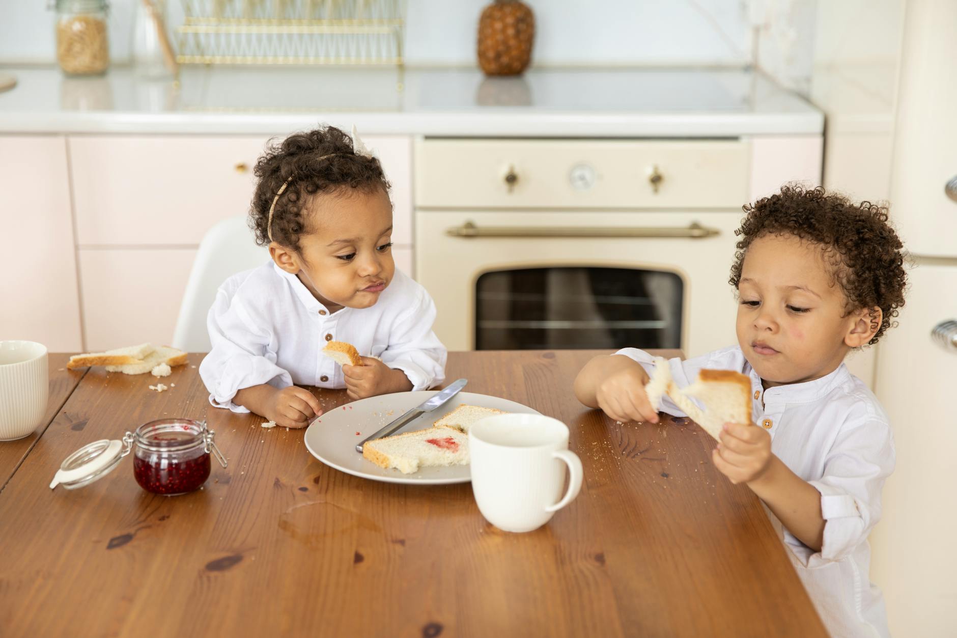 Two young children having breakfast in a bright kitchen, enjoying bread with jam. - teaching toddlers sharing