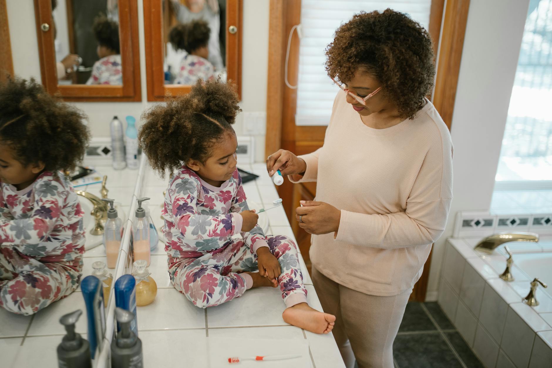 A parent helps a child brush teeth in a bathroom during a morning routine. - toddler self care