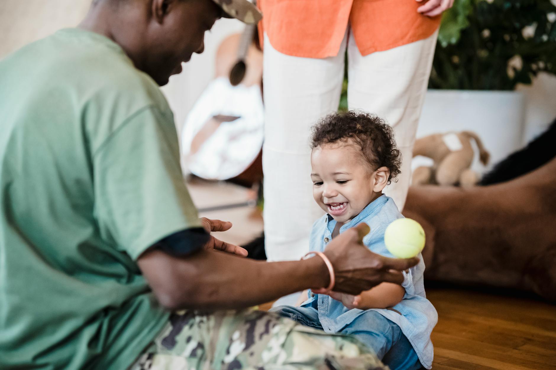 A toddler joyfully plays with a parent holding a tennis ball in a cozy home setting. - toddler sleepaway camp