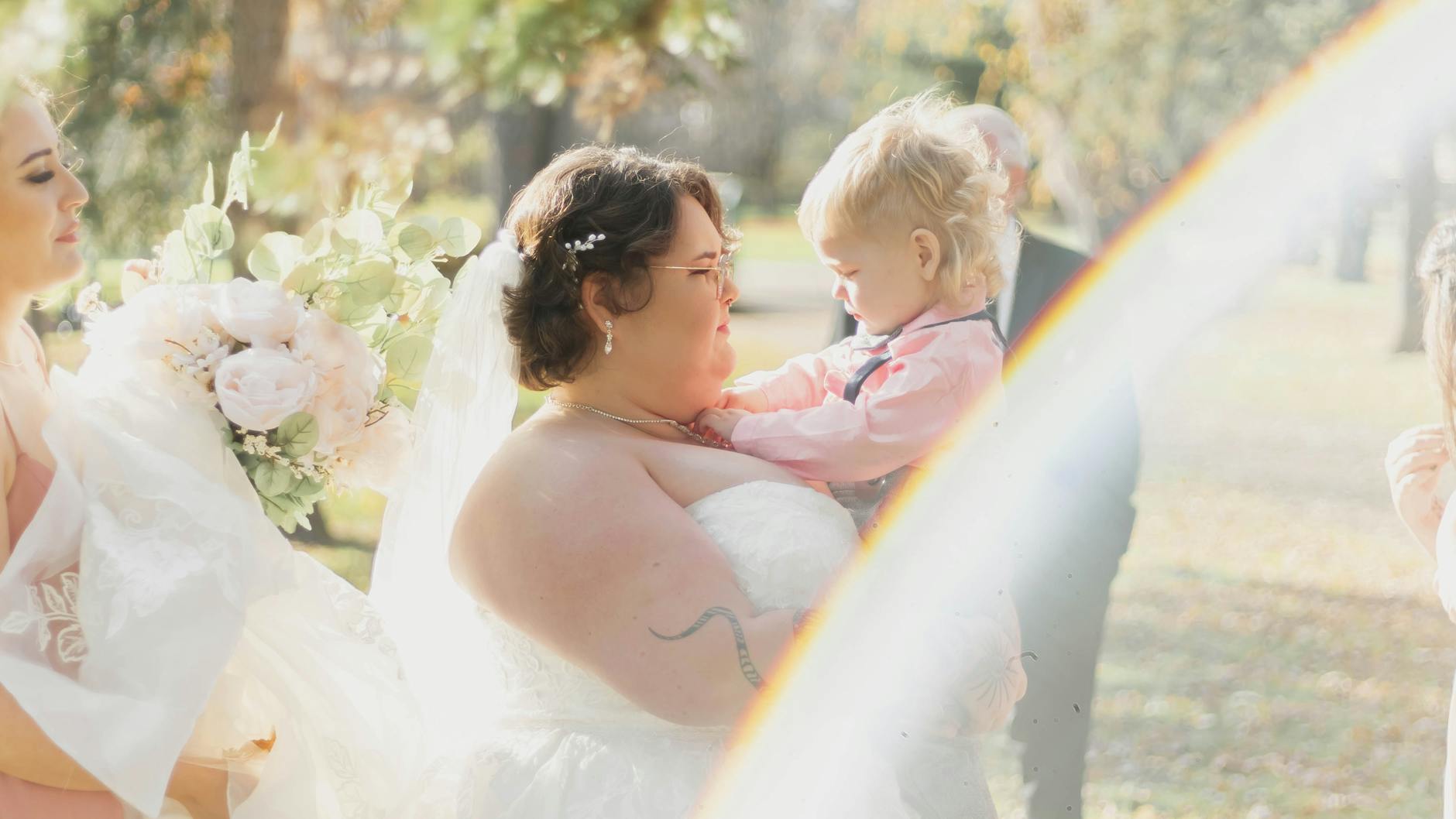 A bride in a white wedding dress holds a child during a sunny outdoor ceremony, surrounded by floral arrangements. - wedding day anxiety